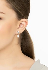 Csepp alakú fülbevalók ezüst színű kerettel, melyek fehér gyöngy csepp alatt egy kerek kristály díszítéssel rendelkeznek, és a fülcimpától lógnak.