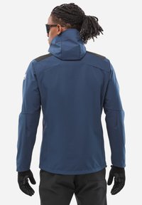 Homme avec des cheveux tressés et des lunettes de soleil, portant une veste à capuche bleue, des gants noirs et un pantalon noir, de dos sur un fond blanc.