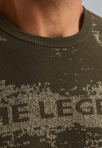 Olijfgroen vest met een gestructureerde breiwijze en een licht patroon, afgewerkt met de opvallende tekst "DE LEGEND" over de borst, ronde halslijn.