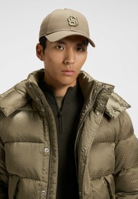 Olijfgroene gewatteerde jas met een ruitpatroon, rits aan de voorkant en drukknoopsluiting. Beige pet met een geborduurd logo op de voorkant.