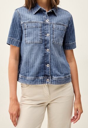 Femme portant une chemise en jean à manches courtes boutonnée avec des rayures verticales et un pantalon beige, montrée des épaules jusqu'à mi-cuisse.