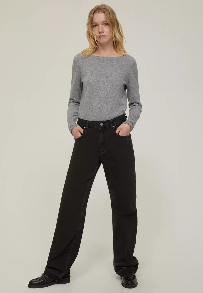 Maglione grigio a maniche lunghe con scollatura a barca abbinato a jeans neri a vita alta e a gamba larga. Il modello sta in piedi con le mani nelle tasche.