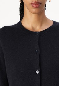 Black knitted cardigan with a round neckline, featuring two button closures and a soft texture. No visible patterns or embellishments.