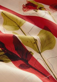 Tissu fleuri présentant un fond crème avec des feuilles rouges et vertes ainsi que des fleurs, mettant en valeur une texture lisse et un contraste de couleurs vif.