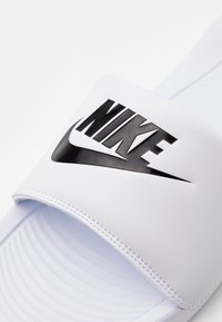 White slide sandal with a wide strap featuring a prominent black Nike logo. The surface is smooth with subtle texture and rounded contours.