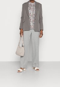Blazer grigio sopra una blusa a fantasia floreale, pantaloni a gamba larga grigio chiaro, borsa beige con manico arricciato e ciabatte beige con tacco blocco.