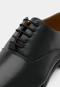 Černá kožená bota s hladkým, lesklým povrchem. Obsahuje šest černých dírek na šněrování a jemný prošívací detail podél okraje.