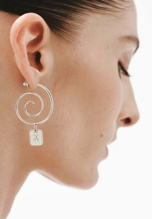Pendientes de plata en forma de espiral con una etiqueta rectangular que presenta una "X" grabada. El diseño es minimalista, con un acabado suave y brillante.