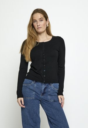 Frau mit langen Haaren, die eine schwarze, geknöpfte Strickjacke und hoch taillierte blaue Jeans trägt, stehend vor einem einfarbigen weißen Hintergrund.