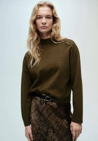 Maglione a collo alto di un verde scuro, con dettagli sottili sulle spalle, abbinato a una gonna di pelle morbidamente rigata con motivo a serpente marrone e una cintura nera.