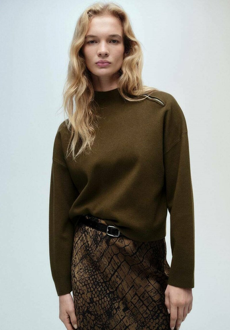 Dunkelgrüner Strickpullover mit hohem Kragen und dezenten Schulterdetails, kombiniert mit einem strukturierten braunen Rock im Schlangenmustermuster und einem schwarzen Gürtel.