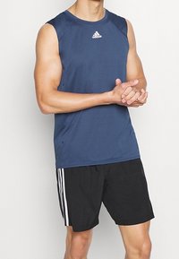 Tmavě modré sportovní tílko bez rukávů s černým lemem a logem Adidas, doplněné černými šortkami s bílými bočními pruhy.