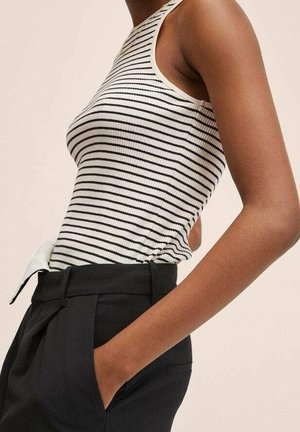 Frau trägt ein weiß-schwarz gestreiftes ärmelloses Top, das in hoch taillierte schwarze Hosen gesteckt ist, mit einer Hand in der Tasche.