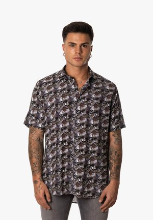 Joven con tatuajes lleva una camisa de manga corta con botones y estampado de calaveras y flores, de pie frente a un fondo liso.