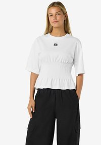 Balta marškinėliai su trumpomis rankovėmis, pasižymintys susiraukšlėjusia juosmens dizainu ir mažuoju juodu logotipu ant krūtinės, derinami su juodomis kargo kelnėmis.