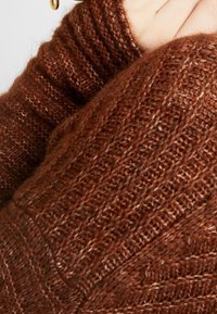 Brauner gestrickter Pullover im Detail, der den strukturierten Stoff mit einem gerippten Muster und einer weichen Oberfläche zeigt. Die Farbe variiert leicht in verschiedenen Brauntönen.