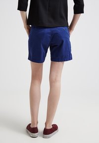 Osoba v modrých šortkách, černém triku s tříčtvrtečními rukávy a vínových slip-on botách, stojící s rukama v kapsách vzadu.