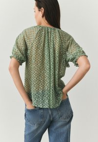 Groene transparante blouse met zwarte stippen, ronde halslijn, korte gerimpelde mouwen en plooien aan de achterkant, gecombineerd met blauwe spijkerbroeken.