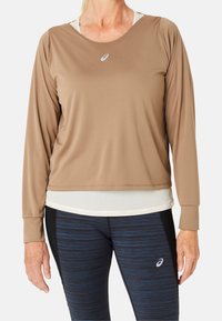 Langärmliges, hellbraunes Sportoberteil aus glattem Stoff, mit rundem Ausschnitt und kleinem Logo auf der Brust. Kombiniert mit dunkel gemusterten Leggings.