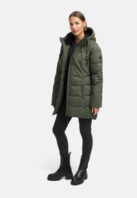 Grön vadderad vinterjacka med en texturerad överdel, huva, svarta accenter och en längre nederdel, parat med svarta skinny jeans och kraftiga svarta stövlar.