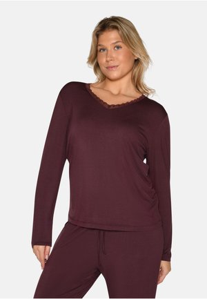 Donna che indossa una maglia a maniche lunghe color viola scuro con scollo a V e bordatura in pizzo e pantaloni abbinati, posando davanti a uno sfondo semplice.