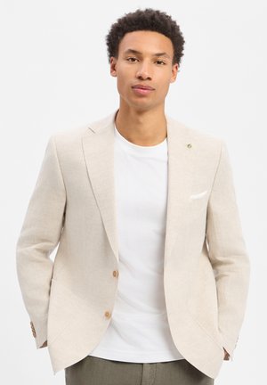 Junger Mann mit lockigem Haar, der einen beigen Blazer über einem weißen T-Shirt trägt, steht mit den Händen in den Taschen vor weißem Hintergrund.