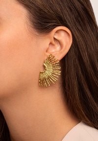 Goldfarbene Ohrringe mit einem Sonnenstrahlen-Design, die verlängerte, strukturierten Strahlen zeigen, die von einer gebogenen Basis ausstrahlen und am Ohr befestigt sind.