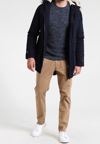 Marinblå parka-jacka med foder i päls, texturerad marinblå tröja, khakifärgade byxor och vita sneakers. Modellen står med armarna längs sidorna på en ljus bakgrund.