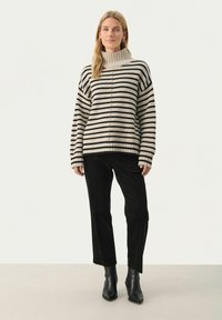 Cremfarbener und schwarzer gestreifter Strickpullover mit hohem Kragen, lockerer Passform und geripptem Saum, kombiniert mit schwarzen Cordhosen und schwarzen Stiefeln mit Absätzen.