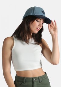 Casquette en denim avec une visière bleu foncé et une calotte bleu clair, ornée d'un logo brodé. Le mannequin porte un crop top blanc côtelé et un pantalon vert.