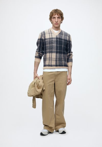 Jeune homme portant un pull à carreaux beige et noir, pantalon large beige, baskets blanches, tenant une veste beige dans une main, debout sur fond blanc.