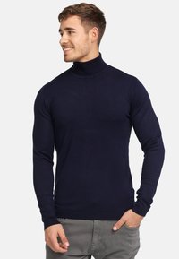 Marine turtleneck trui van glad gebreid materiaal, met lange mouwen en geribde manchetten. Model poseert met handen in de zakken.