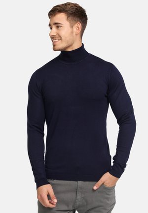 Mørkeblå rullekravesweater lavet af glat strik, med lange ærmer og ribbede manchetter. Model poserer med hænderne i lommerne.