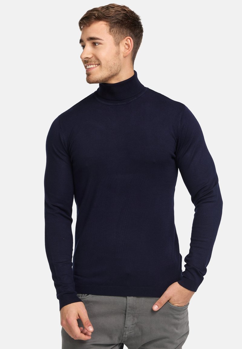 Marine turtleneck trui van glad gebreid materiaal, met lange mouwen en geribde manchetten. Model poseert met handen in de zakken.