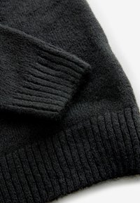 Svart stickad tröja med en texturerad yta, ribbade ärmslut och nederkant, som visar mjukhet och subtil djup i materialet.