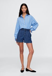 Světle modrá proužkovaná košile s dlouhým rukávem a knoflíky, v kombinaci s vysokým pasem džínovými šortkami a černými plochými botami, stojící před neutrálním pozadím.