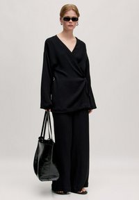Schwarzes, strukturiertes Wickeloberteil kombiniert mit weit geschnittenen schwarzen Hosen. Das Model hält eine glänzende schwarze Tote-Tasche und trägt dunkle Sonnenbrillen. Schlichtes Design.