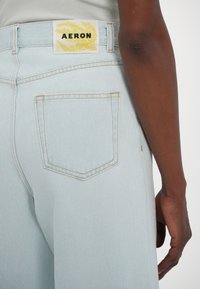 Light blue denim jeans with a straight cut, featuring a yellow label with "AERON" and back pocket stitching in a contrasting color.