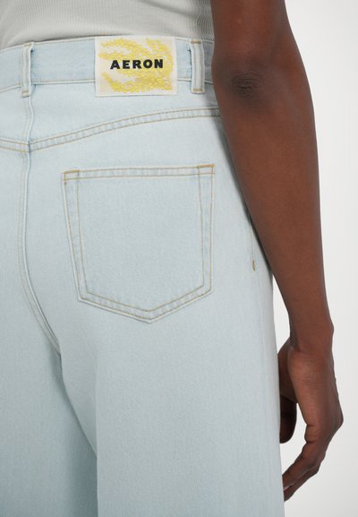 Jeans de denim azul claro com corte reto, apresentando uma etiqueta amarela com "AERON" e costura do bolso traseiro em cor contrastante.