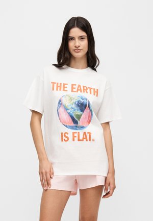 Jonge vrouw met lang donker haar draagt een wit T-shirt waarop de aarde te zien is met een roze bikini topje en de tekst 'DE AARDE IS PLAT.'