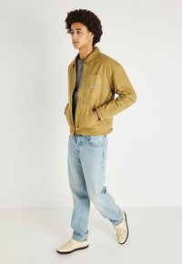 Καφέ καμβάς μπουφάν με τσέπη στο στήθος, σε συνδυασμό με ανοιχτό γαλάζιες τζιν και κρεμ αθλητικά παπούτσια. Το μοντέλο έχει σγουρά μαλλιά και στέκεται σε προφίλ.