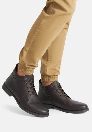 Brązowe skórzane botki za kostkę z okrągłym noskiem, teksturowanym wykończeniem, wiązane na sznurowadła i zapinane na boczny zamek, w połączeniu z beżowymi spodniami z elastycznym mankietem.