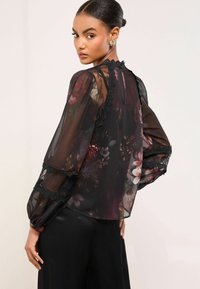 Priehľadná čierna blúzka s kvetinovým vzorom, čipkovými akcentmi, nadýchanými rukávmi a vysokým výstrihom, kontrastujúca s tmavým saténovým spodkom.