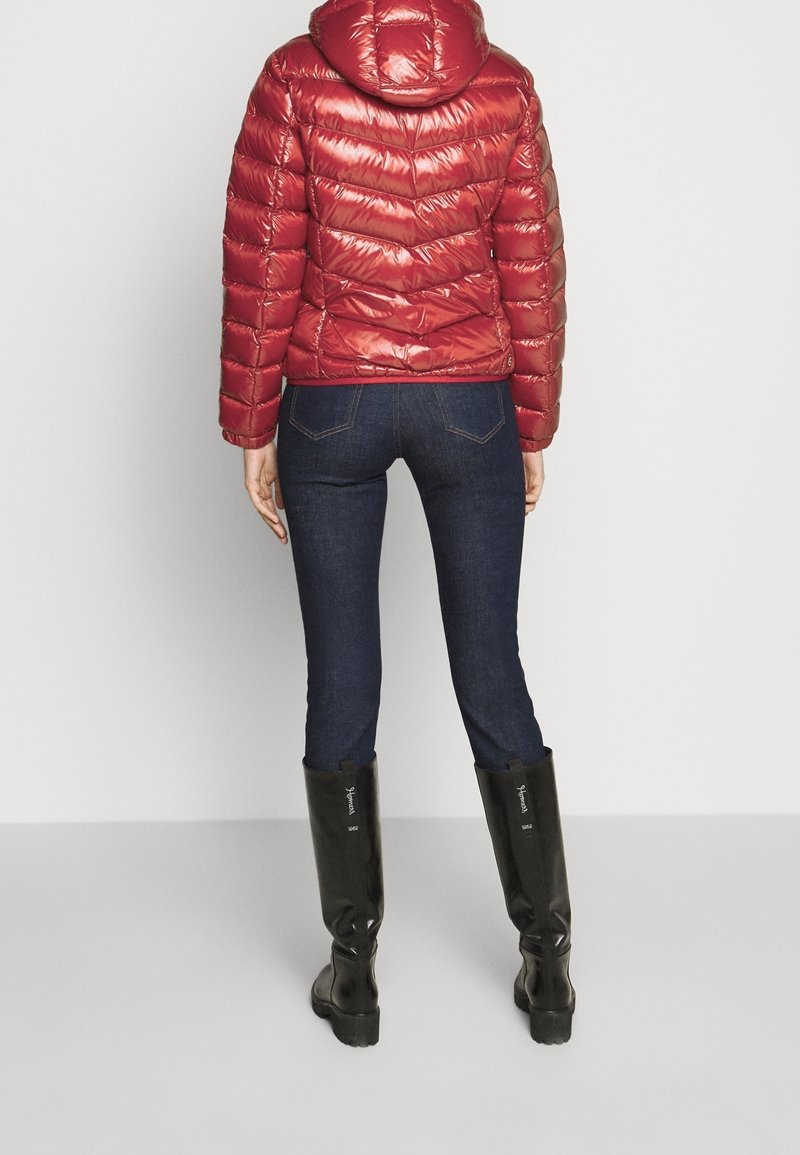 Veste à capuche rouge, rembourrée et brillante, associée à un jean bleu foncé et des bottes noires jusqu'aux genoux. Coutures visibles et détails de rembourrage.