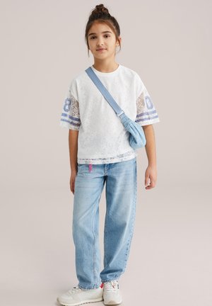 Fehér csipkés póló kék csíkos díszítéssel, világoskék farmerrel és egy kis kék oldaltáskával párosítva; a megjelenést fehér sportcipő egészíti ki.