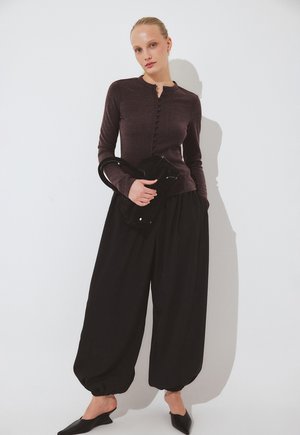 Žena v tmavohnedom zapínanom kardigane, voľných čiernych nohaviciach, v čiernych špicatých topánkach, držiaca čiernu kabelku na bielom pozadí.