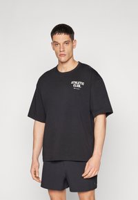T-shirt en coton noir à coupe décontractée, col rond et manches courtes. Présente le texte "ATHLETIC CLUB." en blanc sur la poitrine.