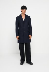 Caban din lână bleumarin cu un design croit, având rever decupat, trei butoane și buzunare laterale, asortat cu pantaloni negri cu picior larg.