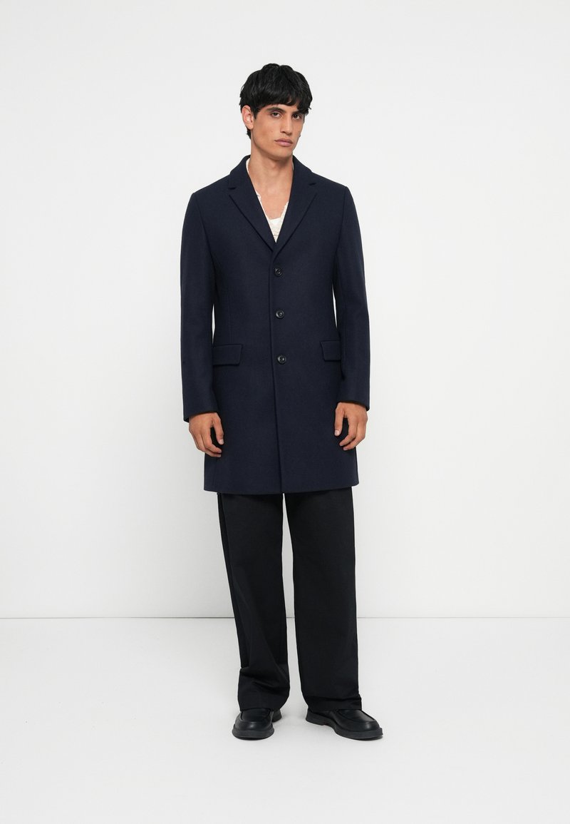 Caban din lână bleumarin cu un design croit, având rever decupat, trei butoane și buzunare laterale, asortat cu pantaloni negri cu picior larg.