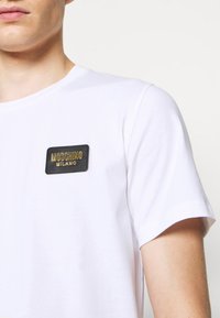 T-shirt blanc à manches courtes avec un patch rectangulaire noir portant le logo Moschino Milano sur le côté supérieur gauche. Texture de tissu douce et lisse.